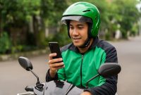 Seorang driver ojek online sedang fokus melihat layar HP terbaik untuk driver ojek online, memantau aplikasi pemesanan dan navigasi saat bekerja di jalanan perkotaan.
